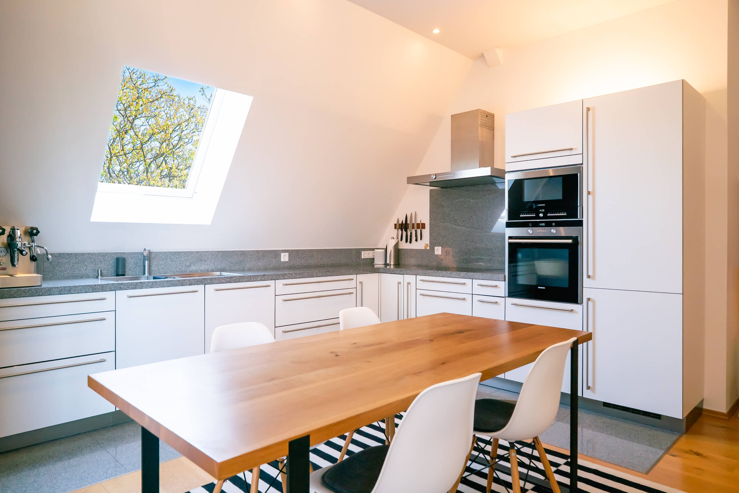 Vista de una cocina moderna en un ático del Quartier 21 de Hamburgo. Mesa de madera con sillas blancas en primer plano, armarios blancos lisos detrás y una vista de los árboles verdes a través de una claraboya.